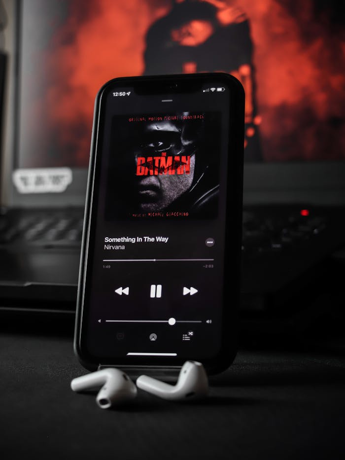 Close-up of a smartphone displaying a music player app with AirPods nearby, featuring Batman album art.